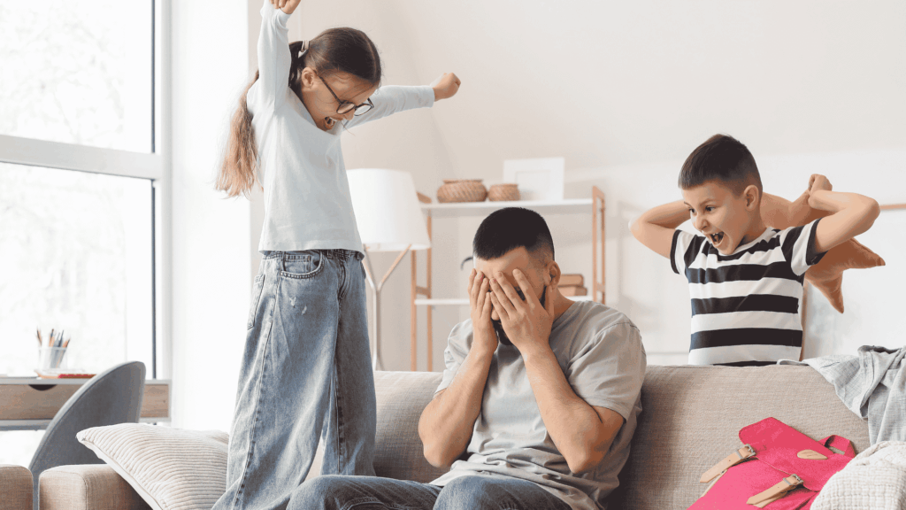 An adult sits on a sofa with hands covering the face while two children stand nearby in a living room. One child has arms raised, and the other is holding a stuffed toy. The scene depicts the challenges of balancing parenting responsibilities and household demands in a busy home environment.